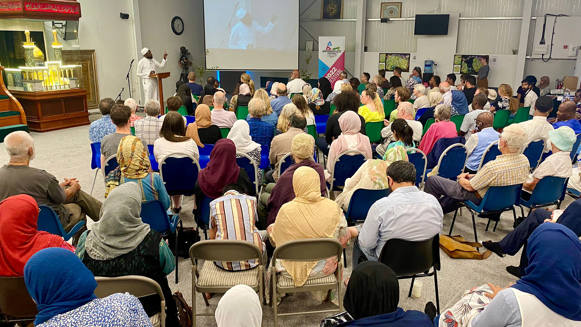 More than 120 people of all ages and faiths sitting down listening to an imam speak